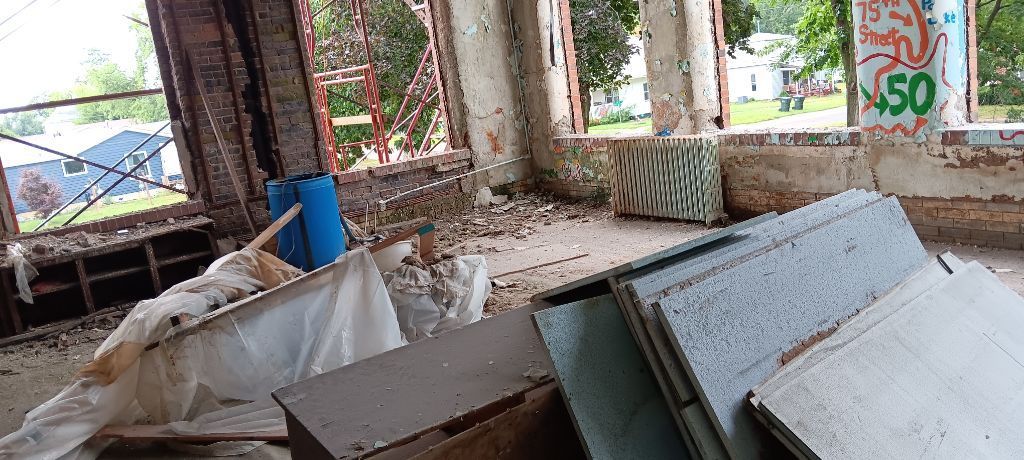 Abandoned room with broken windows, debris, and leaning panels near a graffiti-covered wall.