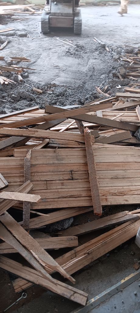 Pile of broken wooden pallets beside a wet, ashy ground with a forklift in the background