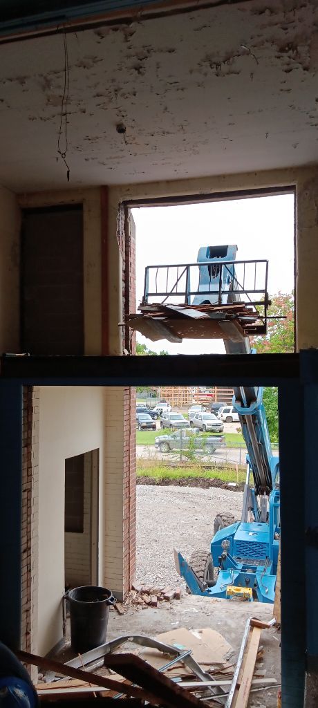Interior view through a doorway to a blue excavator outside a partially demolished building.