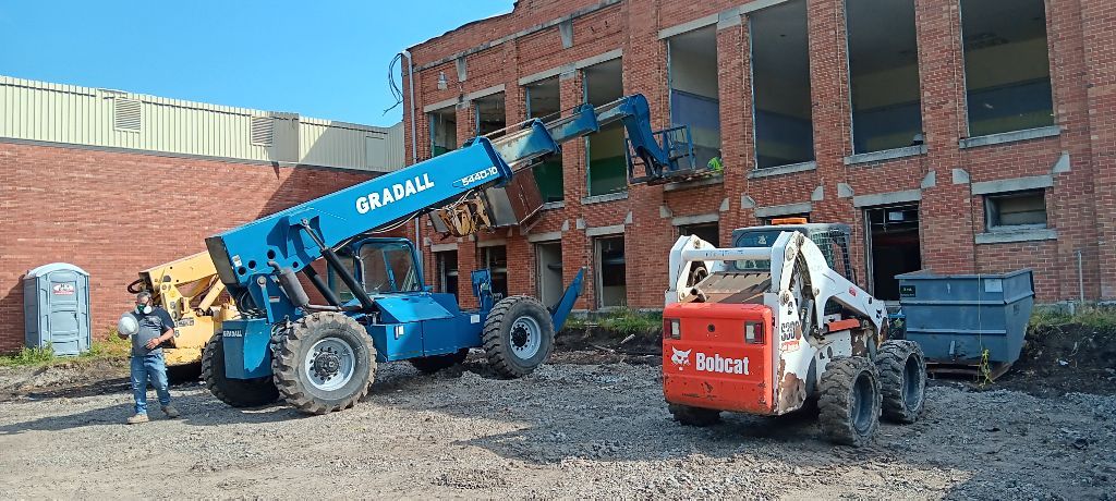 Blue and white construction lifts beside a brick building with dumpsters on a gravel lot