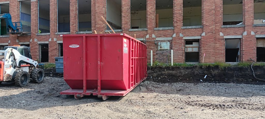 Red construction dumpster beside a brick building with a skid steer in the background