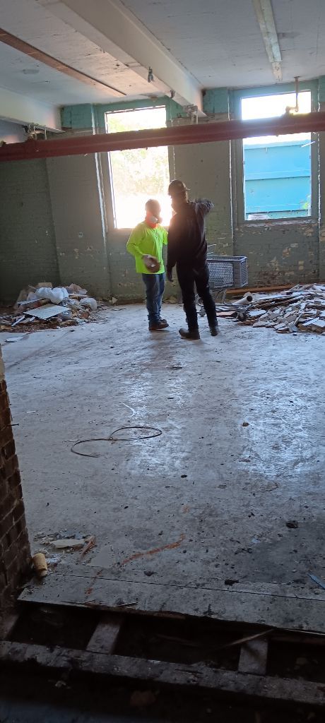 Two people standing in a damaged, debris-filled room with light coming through broken windows.