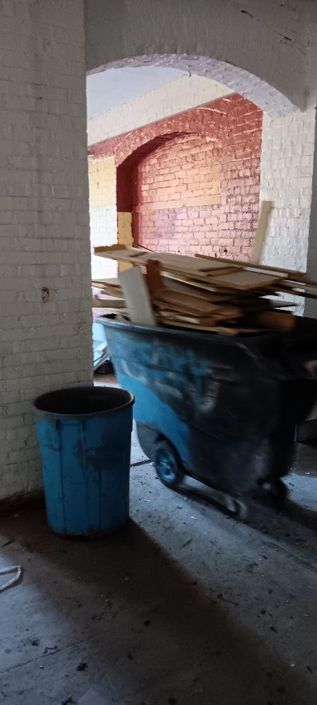 Blue trash bin beside a wheelbarrow full of wood in a concrete archway hallway.
