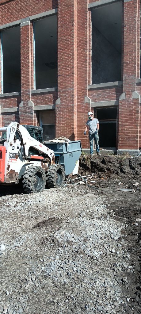 Construction workers and a bobcat loader beside a brick building under renovation