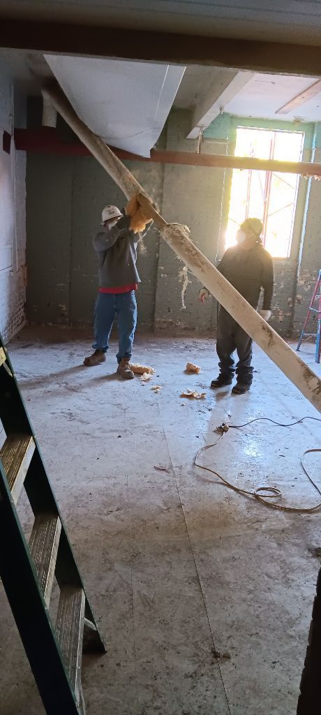Two workers in a dusty room lifting a long beam near a bright window during renovation