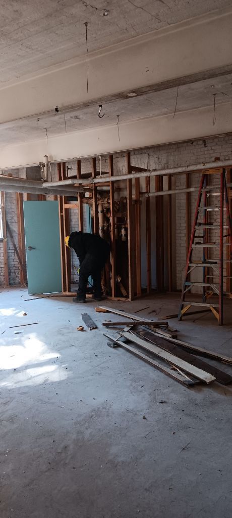 Construction site interior with exposed framing, ladders, debris, and a worker near a blue door