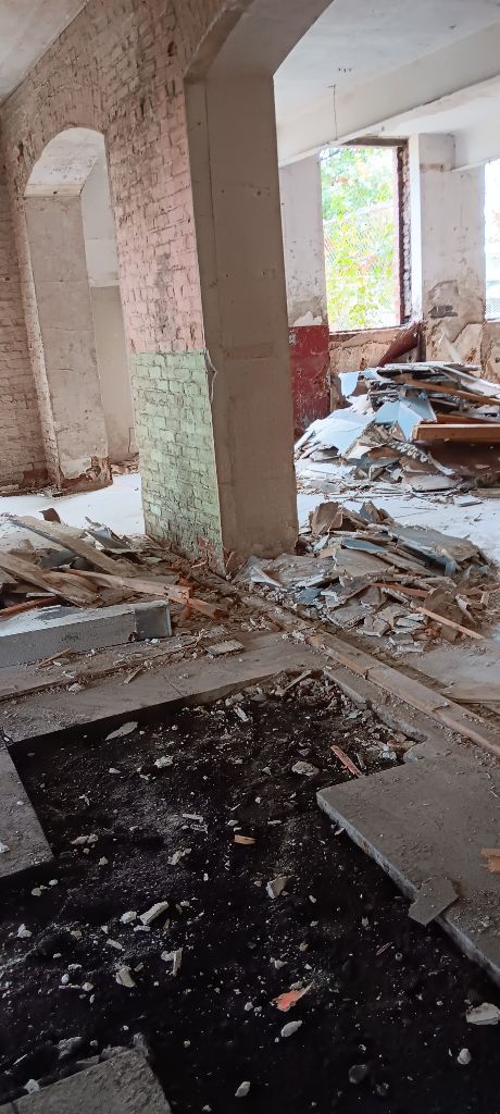 Damaged, abandoned building interior with debris, broken floors, and exposed concrete pillars.