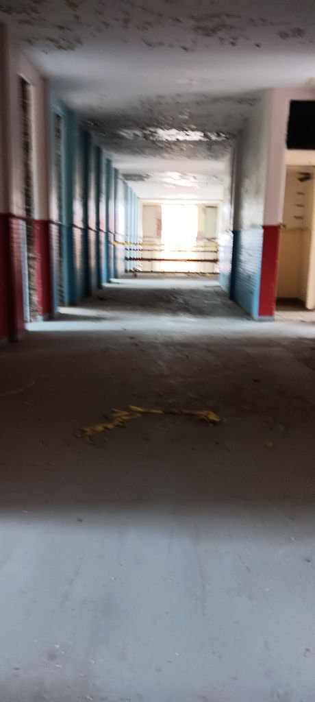 Long, dim abandoned hallway with peeling walls and red-painted columns leading to a bright doorway.