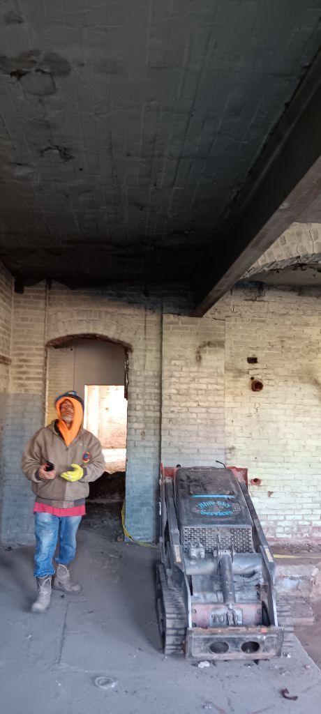 Person in a hooded jacket standing in a dim unfinished room beside stacked bricks and a pallet jack.