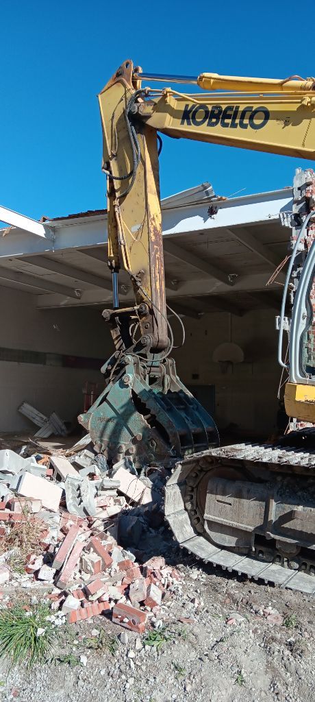 Yellow Kobelco excavator demolishing rubble beside a building under a blue sky