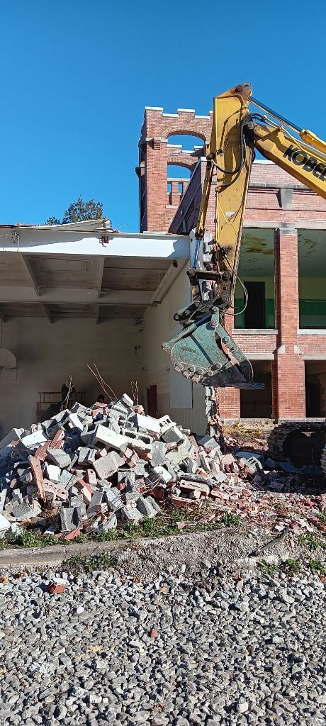 Yellow excavator demolishing a brick building, with rubble piled in front.