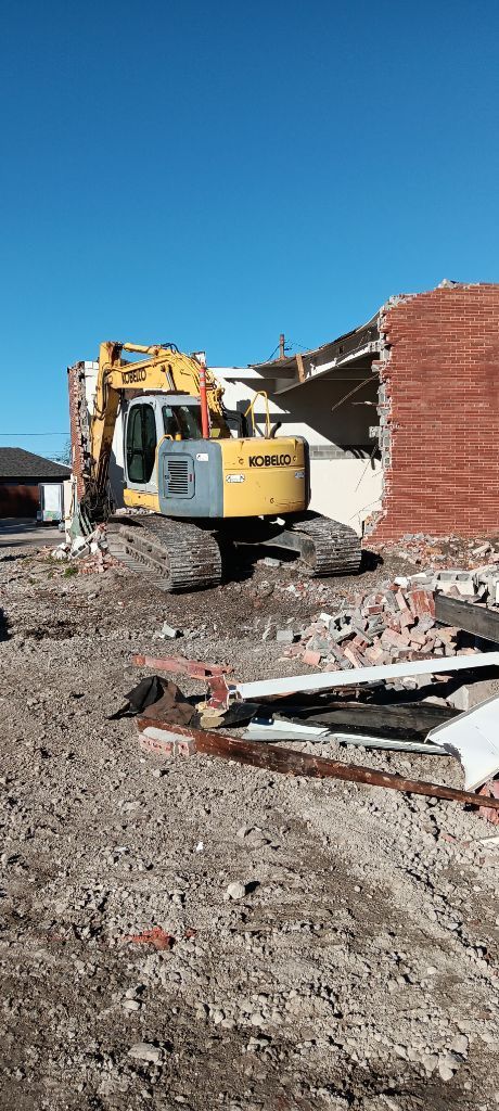 Yellow excavator demolishing a brick building amid rubble under a clear blue sky
