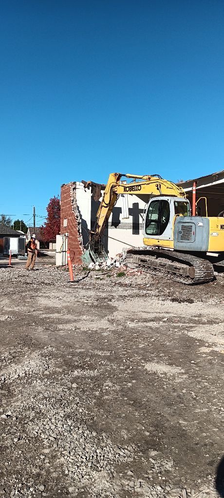 Construction site with yellow excavator beside a partially demolished brick wall under a clear blue sky
