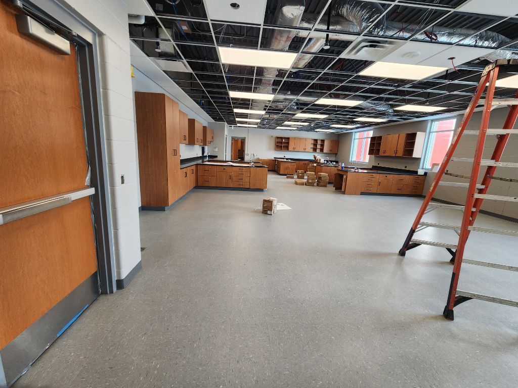 Empty school cafeteria or classroom with tables, gray floor, and a red ladder under a ceiling under repair