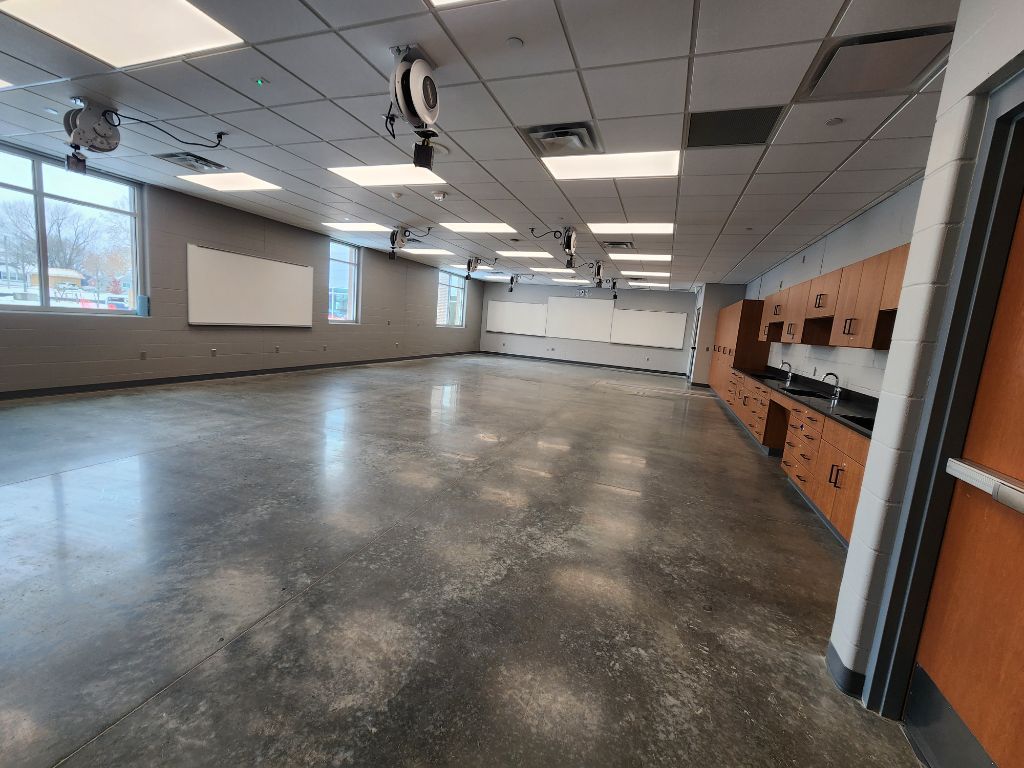 Empty classroom with polished floor, projector screen, windows, and orange storage cabinets along one wall