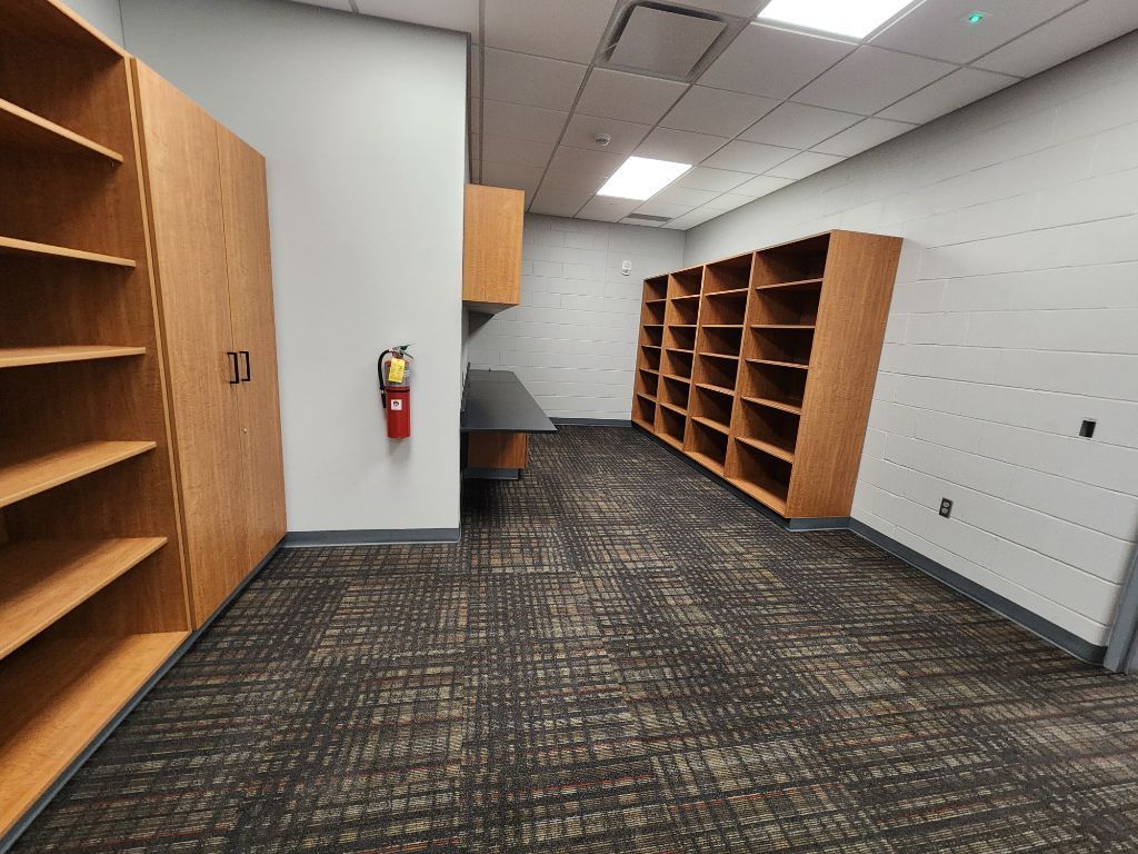 Empty office room with wooden shelving, carpeted floor, and a fire extinguisher on a gray wall.
