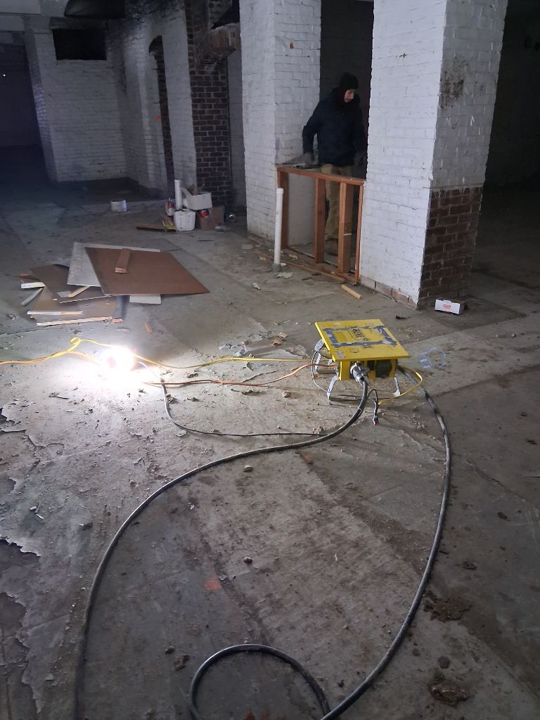 Dim room under construction with debris on the floor, a yellow tool in the foreground, and a person by a table.