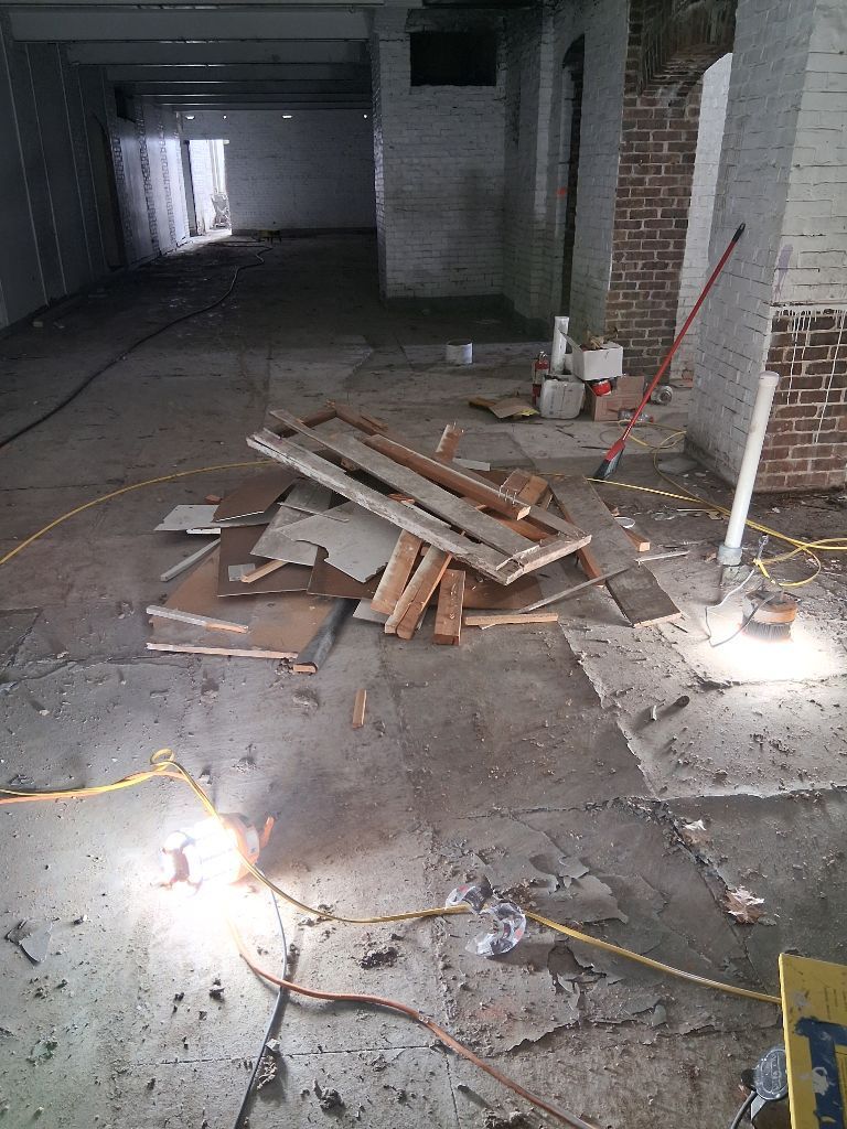 Dim unfinished room with debris pile, exposed brick columns, and scattered cables on a concrete floor.