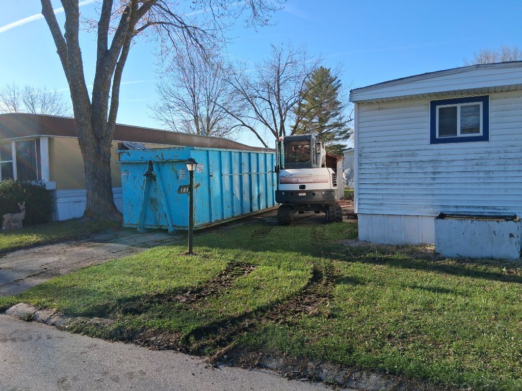 Backyard with blue dumpster beside a white mobile home and parked pickup truck in bright daylight