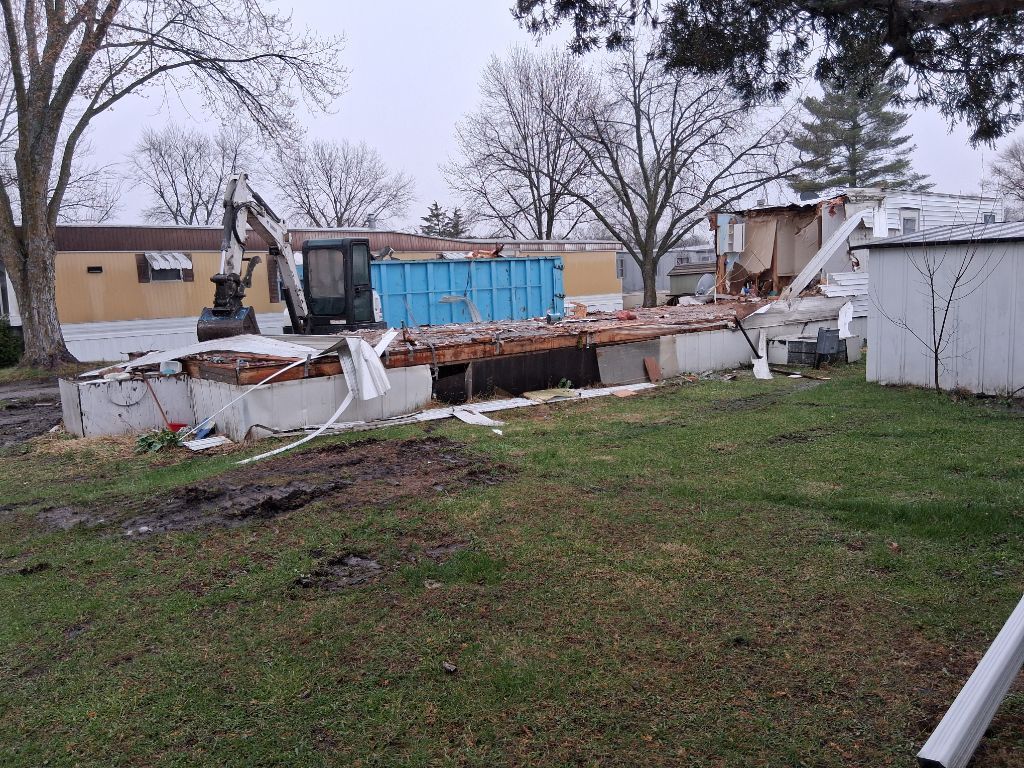 Collapsed building with debris, an excavator, and a blue dumpster in a yard