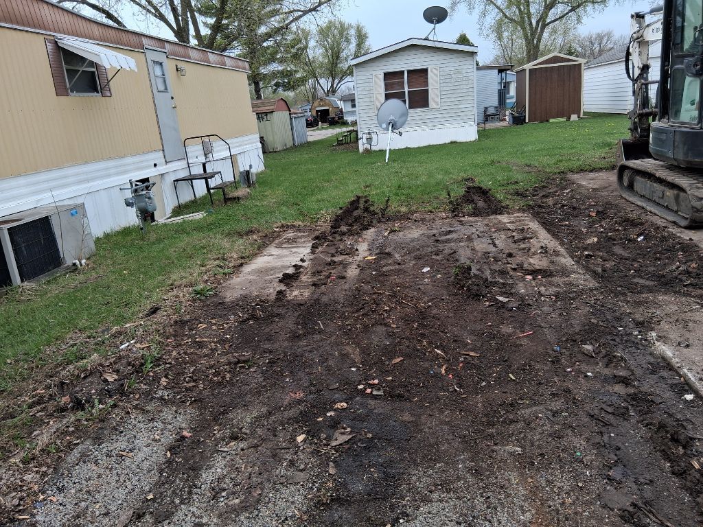 Dirt lot beside a mobile home, with a shed and a small house in the background.