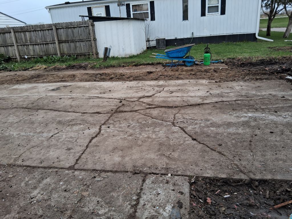 Cracked concrete driveway beside a fenced backyard with a white house and scattered yard tools