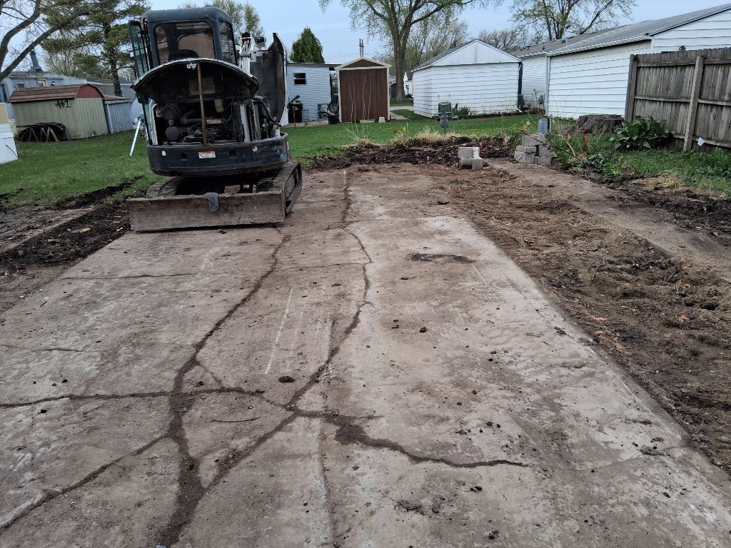 Cracked concrete driveway beside a parked black vehicle and white garage buildings in a backyard.