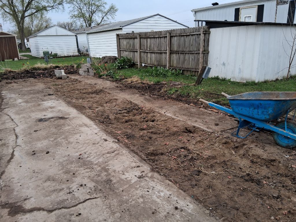 Backyard with a concrete path, dug dirt trench, wooden fence, shed, and blue wheelbarrow.
