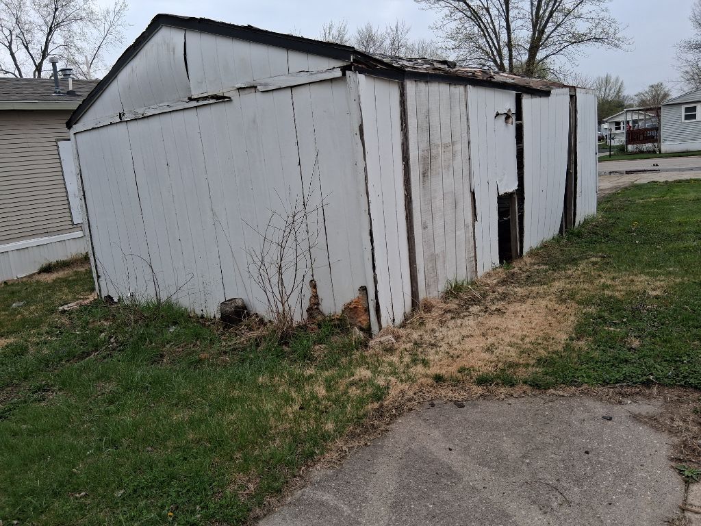 White weathered shed with peeling paint and a slanted roof beside a driveway and grass.