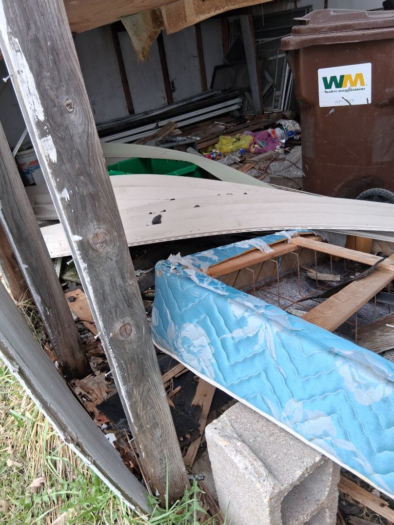 Broken fence and scattered debris in a yard beside a brown WM trash bin.