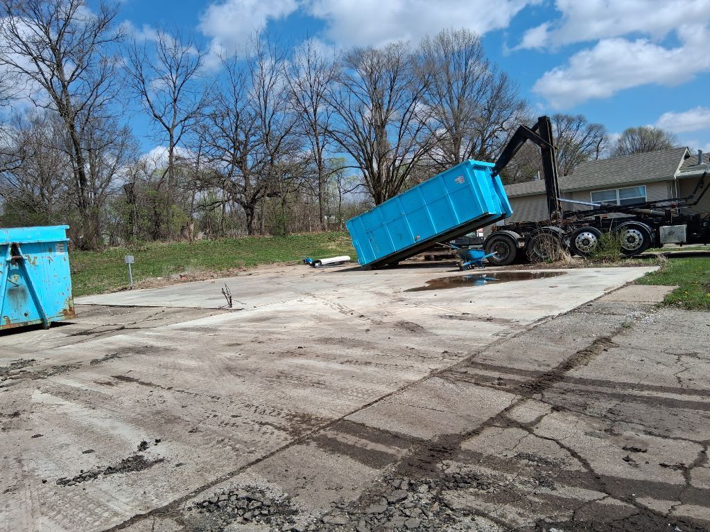 Blue dumpster being hoisted by a truck crane on a concrete lot under a clear sky