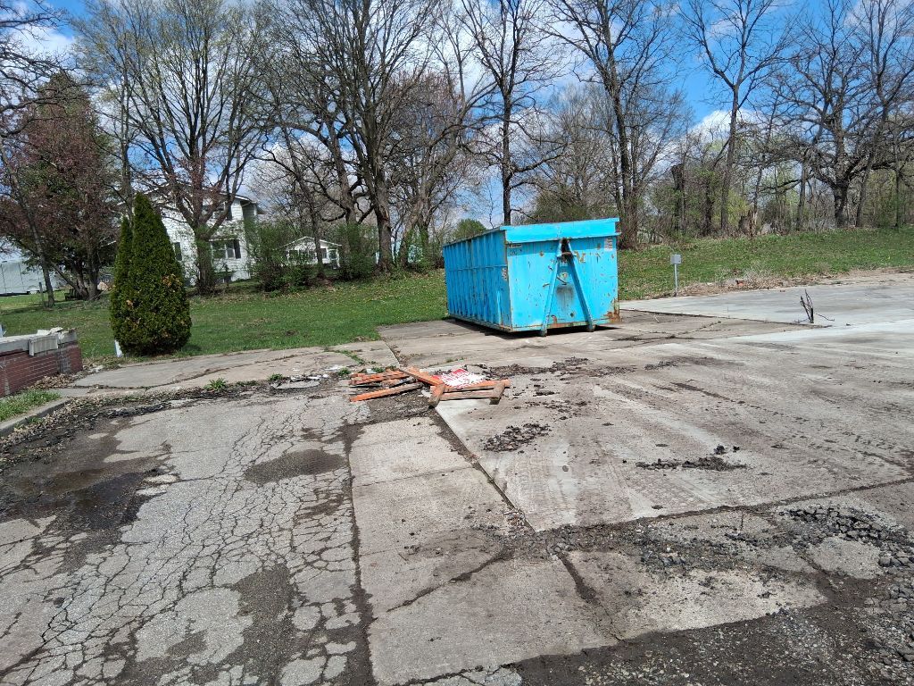 Blue dumpster on cracked concrete lot with scattered debris and trees in the background
