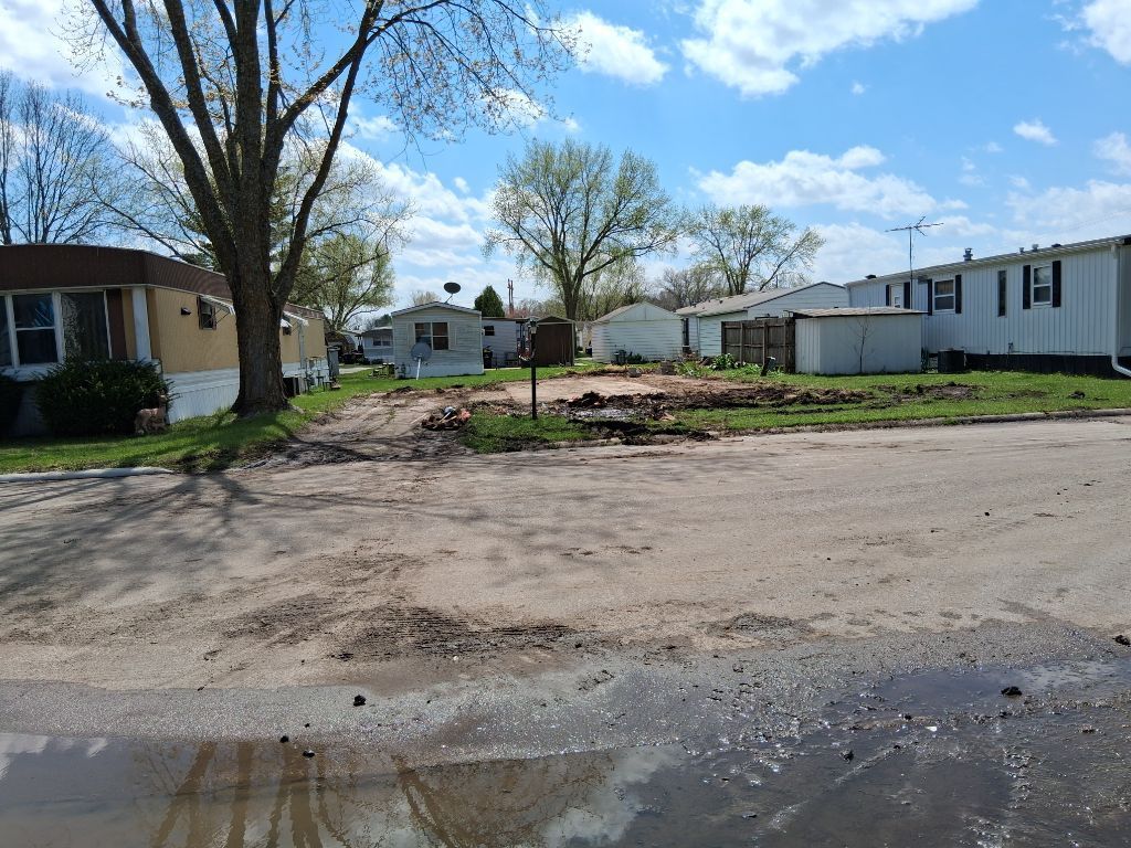 Empty dirt lot between mobile homes under a blue sky with trees and a muddy puddle in the foreground