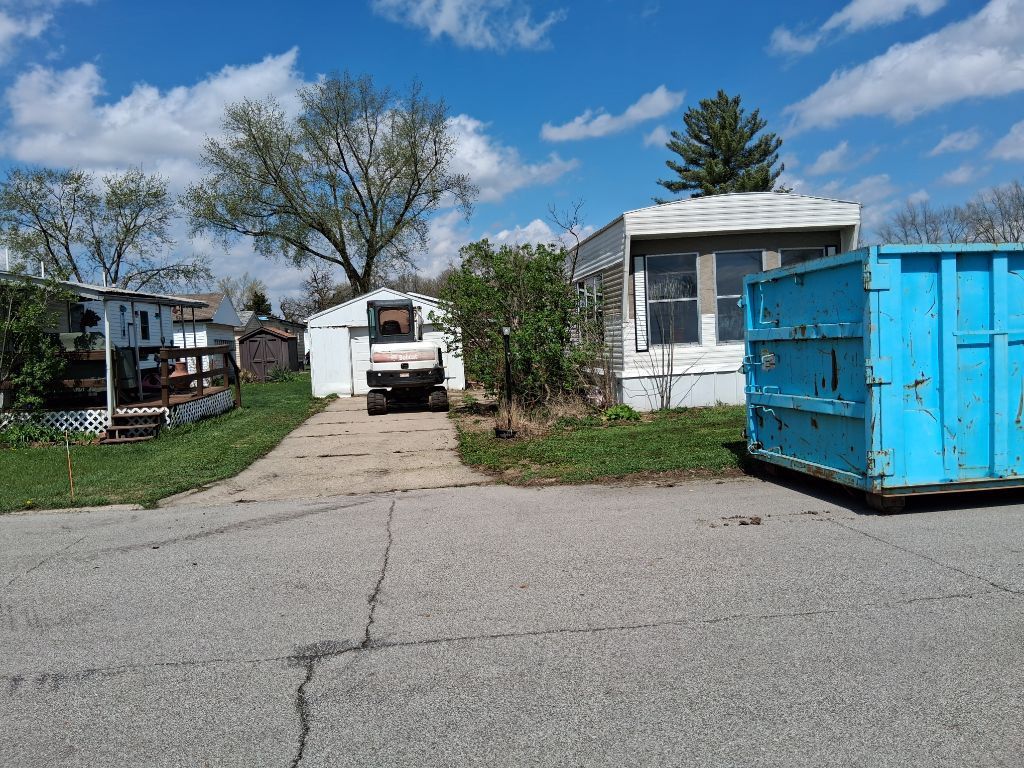Residential street with white houses, parked truck, and a large blue dumpster under a partly cloudy sky