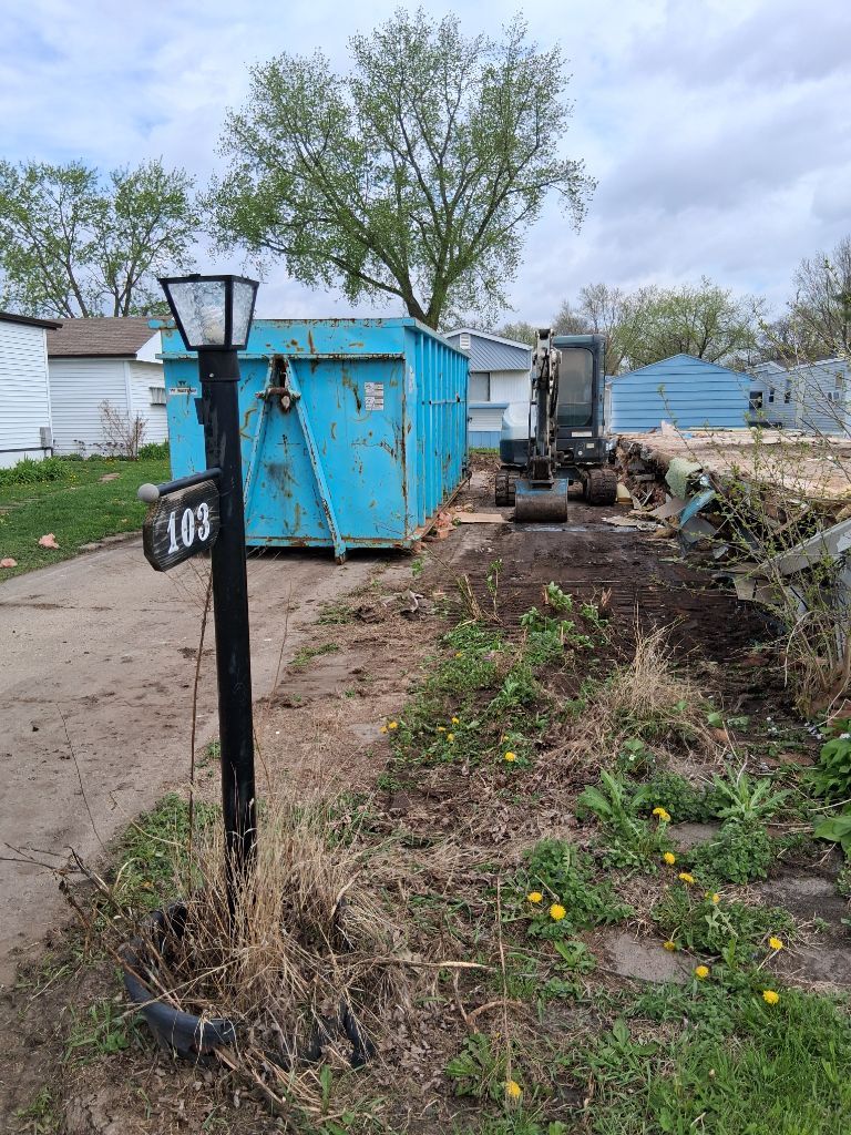 Backyard with blue dumpster, small excavator, and a black lamp post along a muddy path