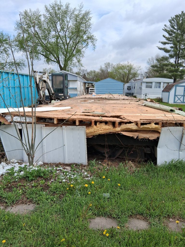 Partially collapsed wooden shed with exposed framing in a grassy yard, surrounded by trees and trailers