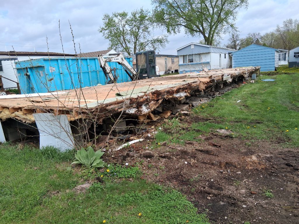 Collapsed wooden platform beside blue sheds on a grassy lot after storm damage