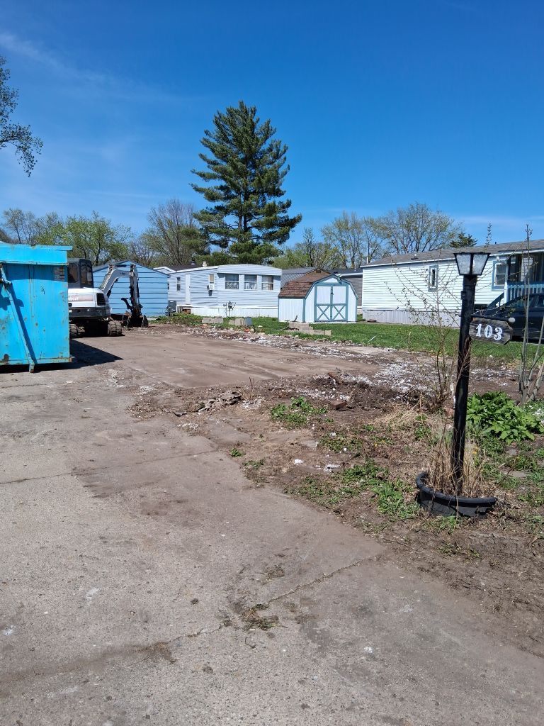 Rural driveway beside a blue dumpster, white mobile homes, and a tall evergreen tree under a clear sky