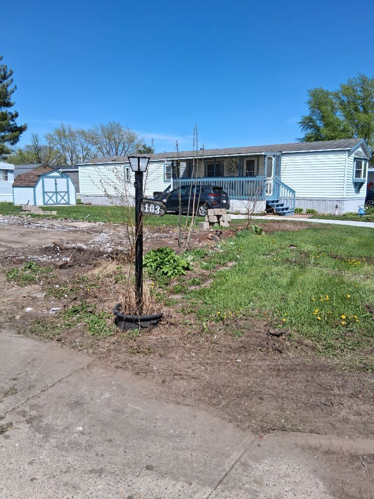 Small white mobile home with porch in a sunny yard, viewed from a dirt driveway under a blue sky.