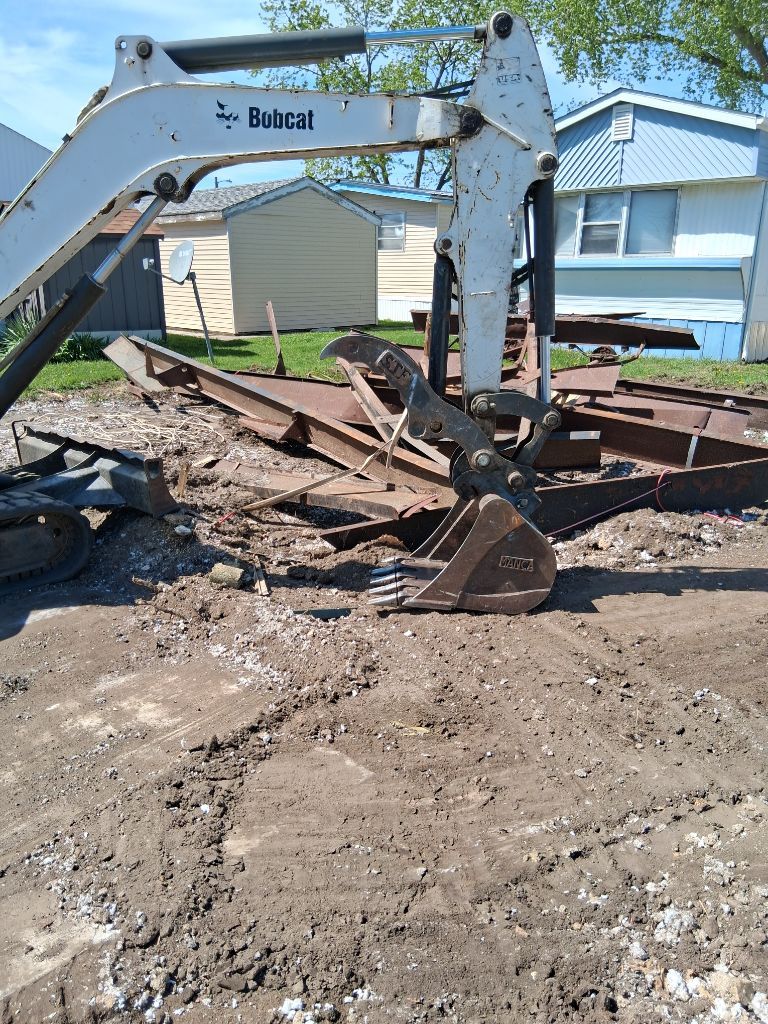Excavator digging up debris in a muddy residential yard beside a house