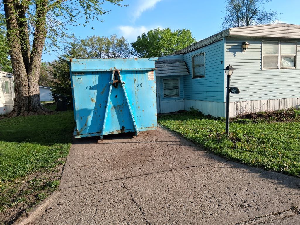 Blue dumpster blocking a driveway beside a light blue house on a sunny day