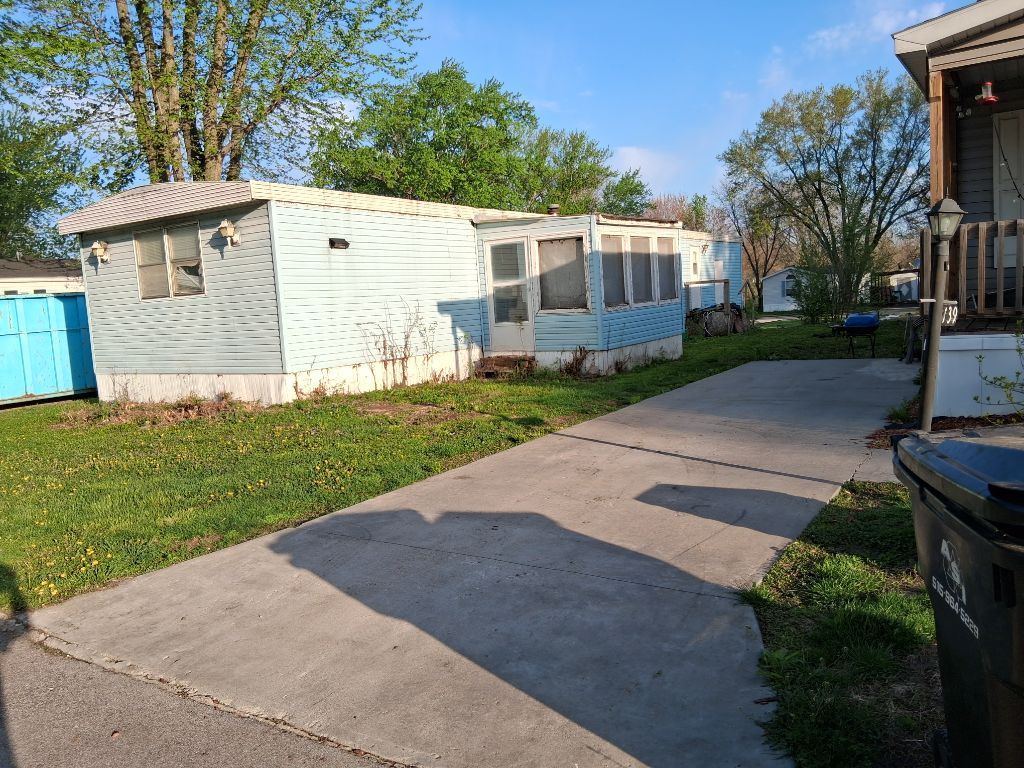 White mobile home beside a concrete driveway, with trees and a blue shed in the yard.