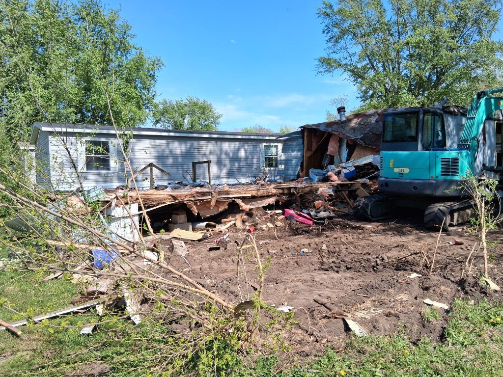 Flood-damaged yard with a damaged blue bus beside a white trailer and scattered debris under trees