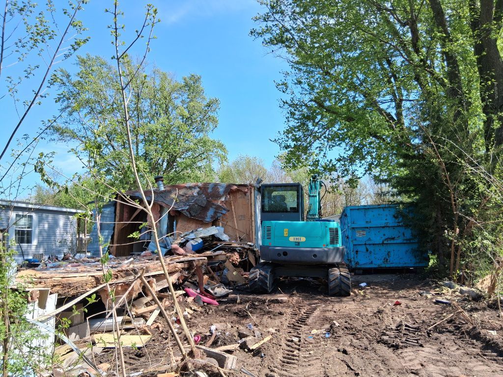 Blue excavator demolishing a small structure in a muddy yard with debris and trees nearby