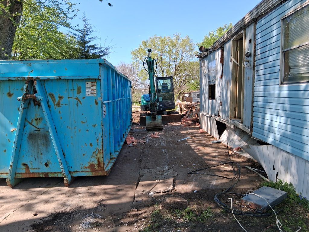 Blue dumpster beside a house with a backhoe in a narrow dirt alley