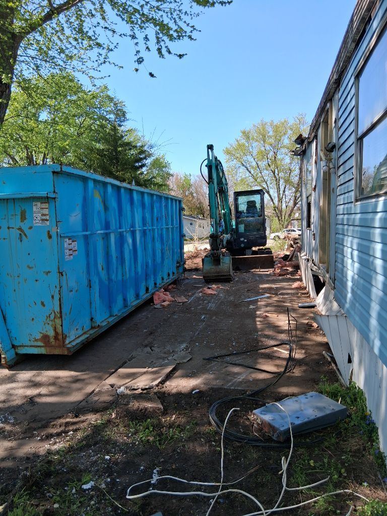 Blue dumpster and excavator in a narrow yard beside a white building, with debris on the ground.