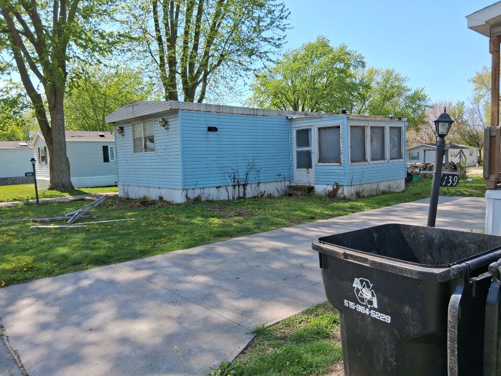Small blue house with white trim beside a driveway, with a black trash bin in the foreground.