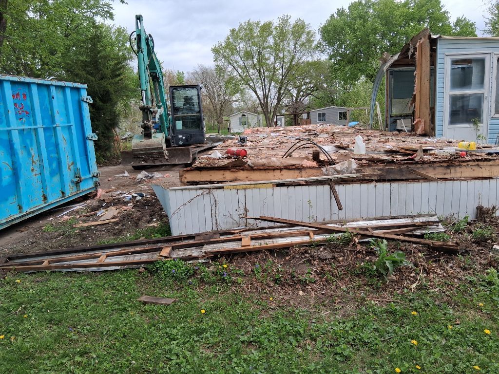 Construction site with excavator, blue dumpster, and debris beside a small white building