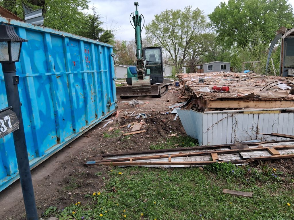 Demolition site with blue dumpster, excavator, and rubble beside a house