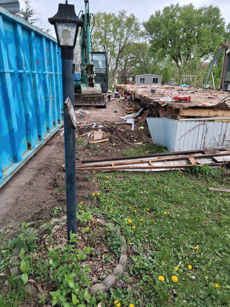 Construction site beside a blue container wall, with a lamp post, dirt trench, and scattered debris.